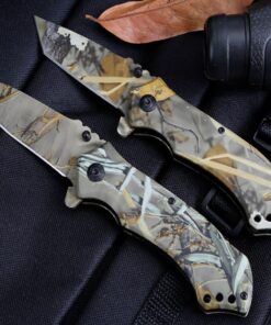 Two camouflage locking knives lying next to a leaf, against a black background.