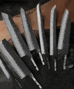 Seven Damascus steel kitchen knife blanks of differing design, displayed against a long wooden chopping board.